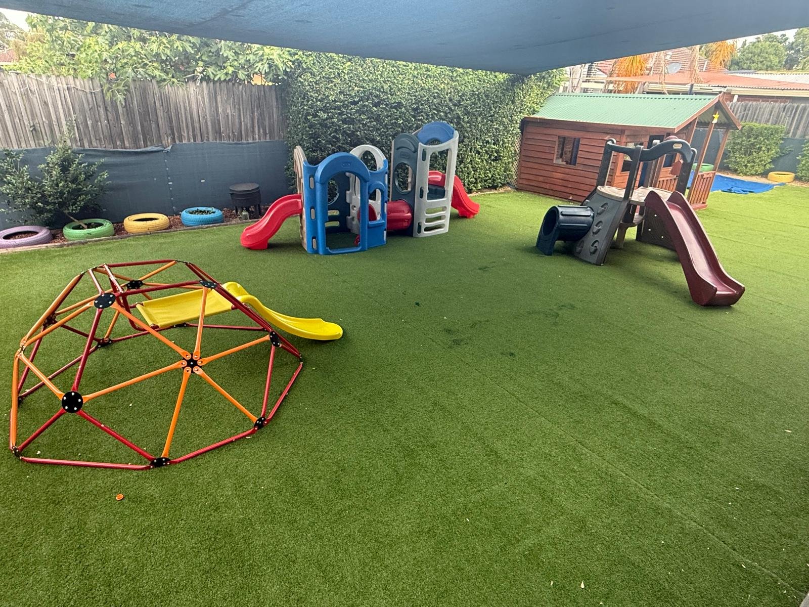 Children's outdoor play area with green artificial turf, multiple small slides, climbing structures, a small wooden playhouse, and colorful tires along the fence.