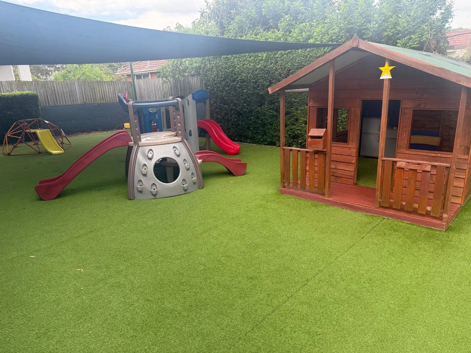 Children's backyard with a small wooden playhouse, a plastic slide and climbing structure, a yellow slide on a climbing dome, and a shaded backyard area with artificial grass.