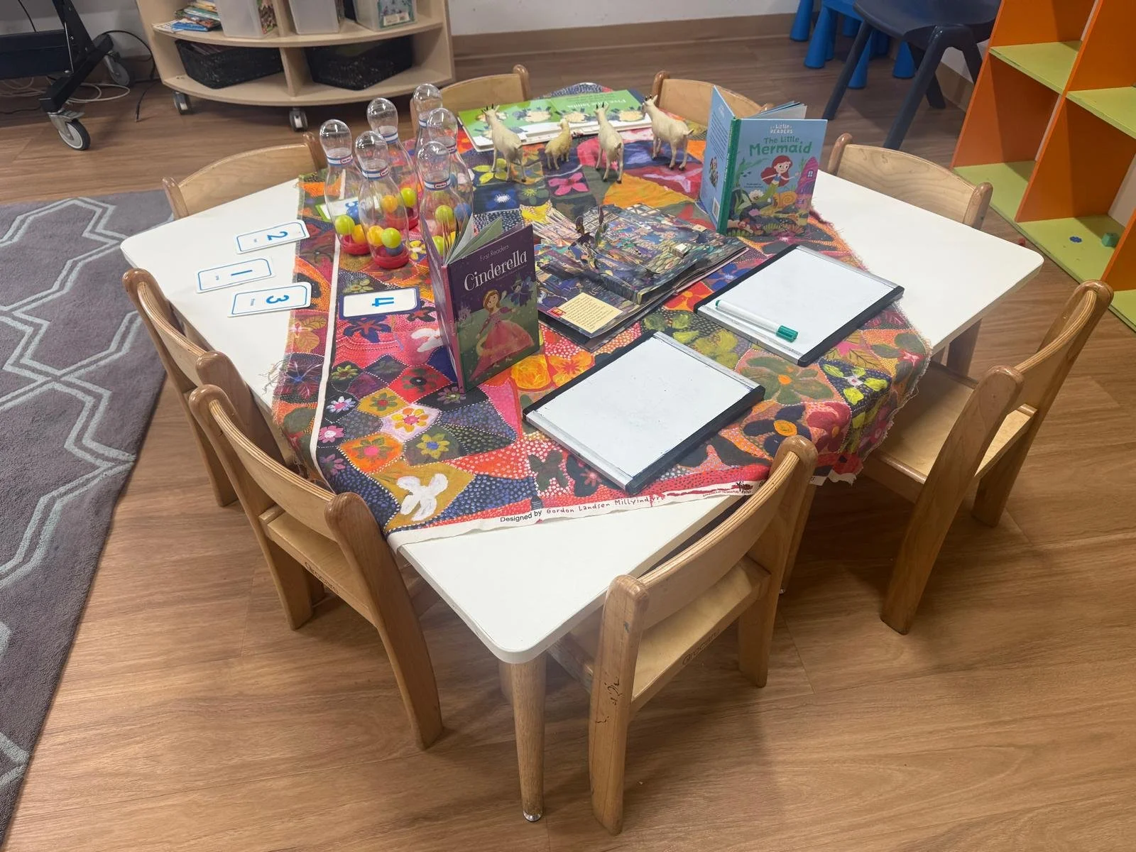 A round table with a colorful cloth, children's books, mini bowling pins with colorful balls, and small animal figurines in a classroom setting.