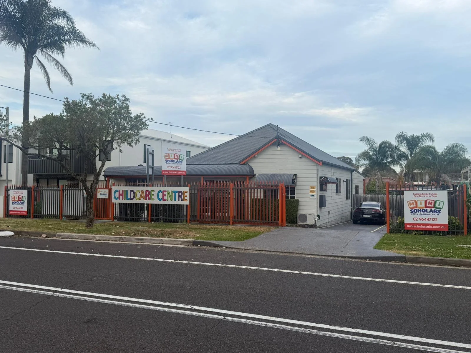 A childcare center with a large sign reading 'Childcare Centre,' surrounded by a red fence. There are smaller signs with the logo 'MINI SCHOLARS' and contact information. The building is white with a gray roof, and there are palm trees and a car park