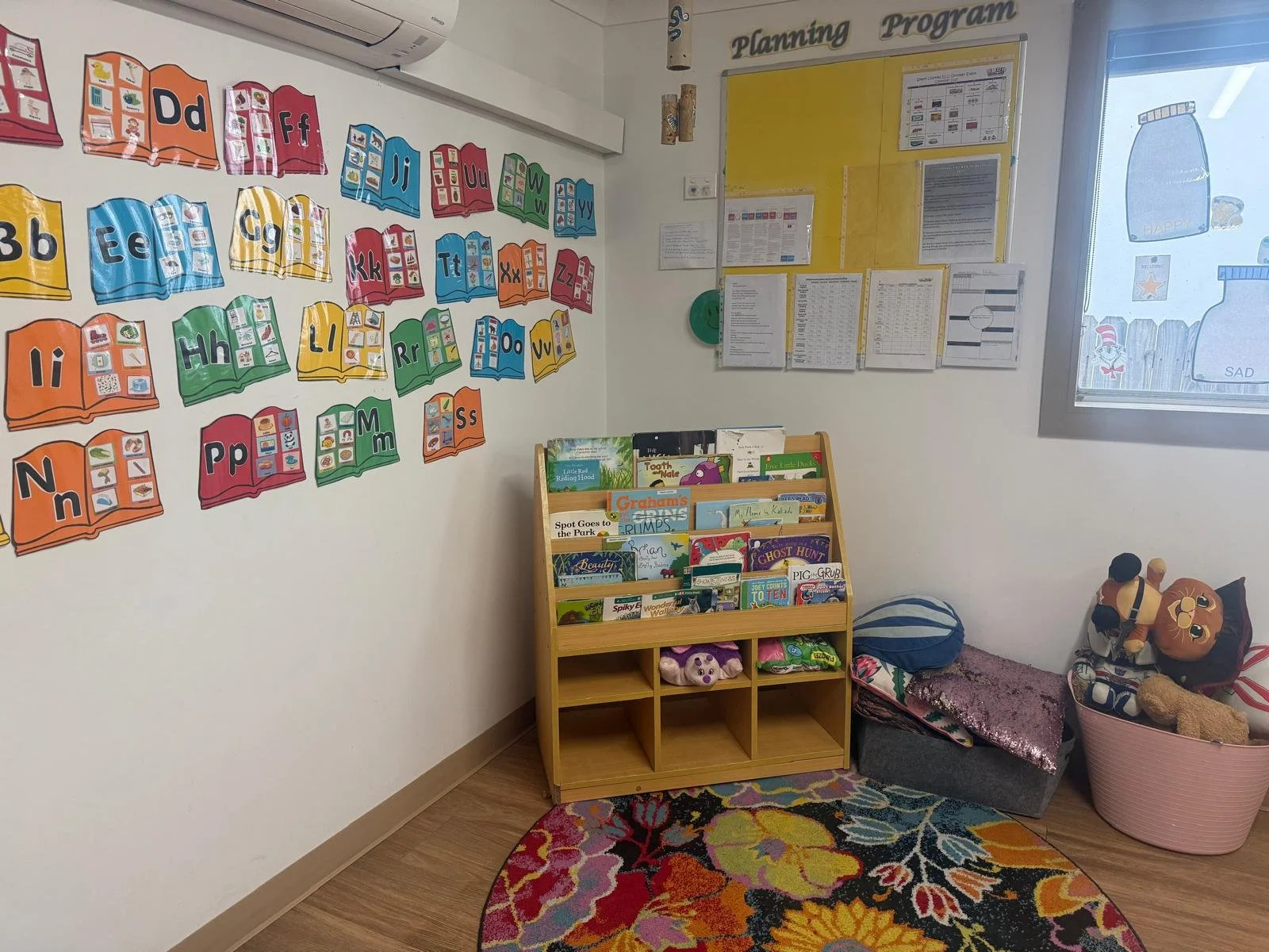 Colorful ABC alphabet bulletin board on wall with open book illustrations, a yellow planning chart, a corner bookshelf filled with children's books, a floral rug, and a window with playful decals showing crayons labeled 'HAPPY' and 'SAD' inside a cla