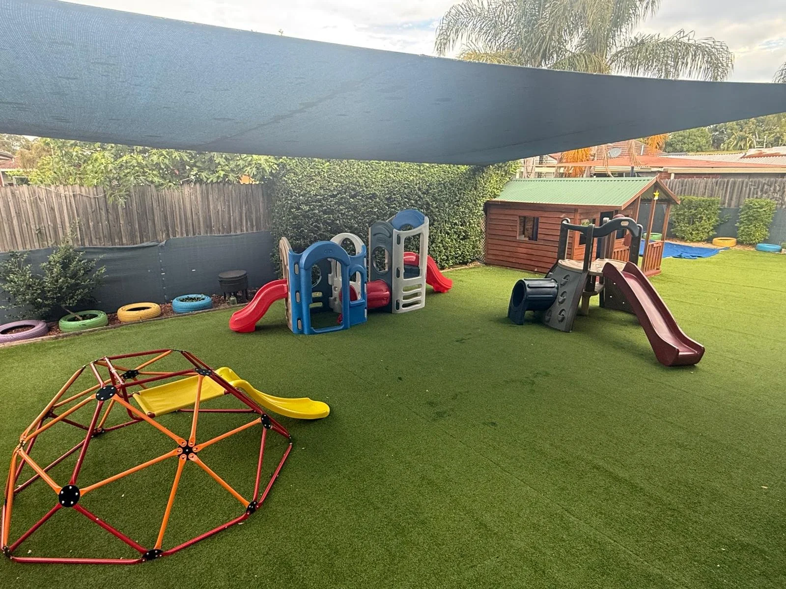 Backyard play area with small slides, a playhouse, climbing equipment, a yellow slide integrated into a climbing dome, and a grassy surface with a shade sail overhead.