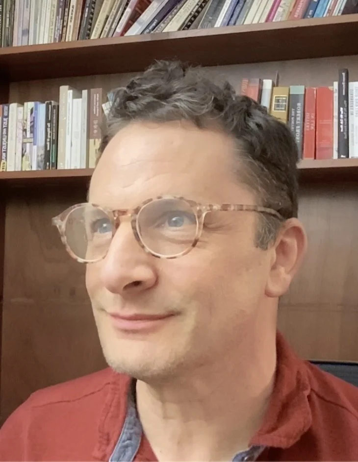 A man with glasses and short dark, slightly curly hair, wearing a red jacket, smiling in front of a bookshelf filled with books.