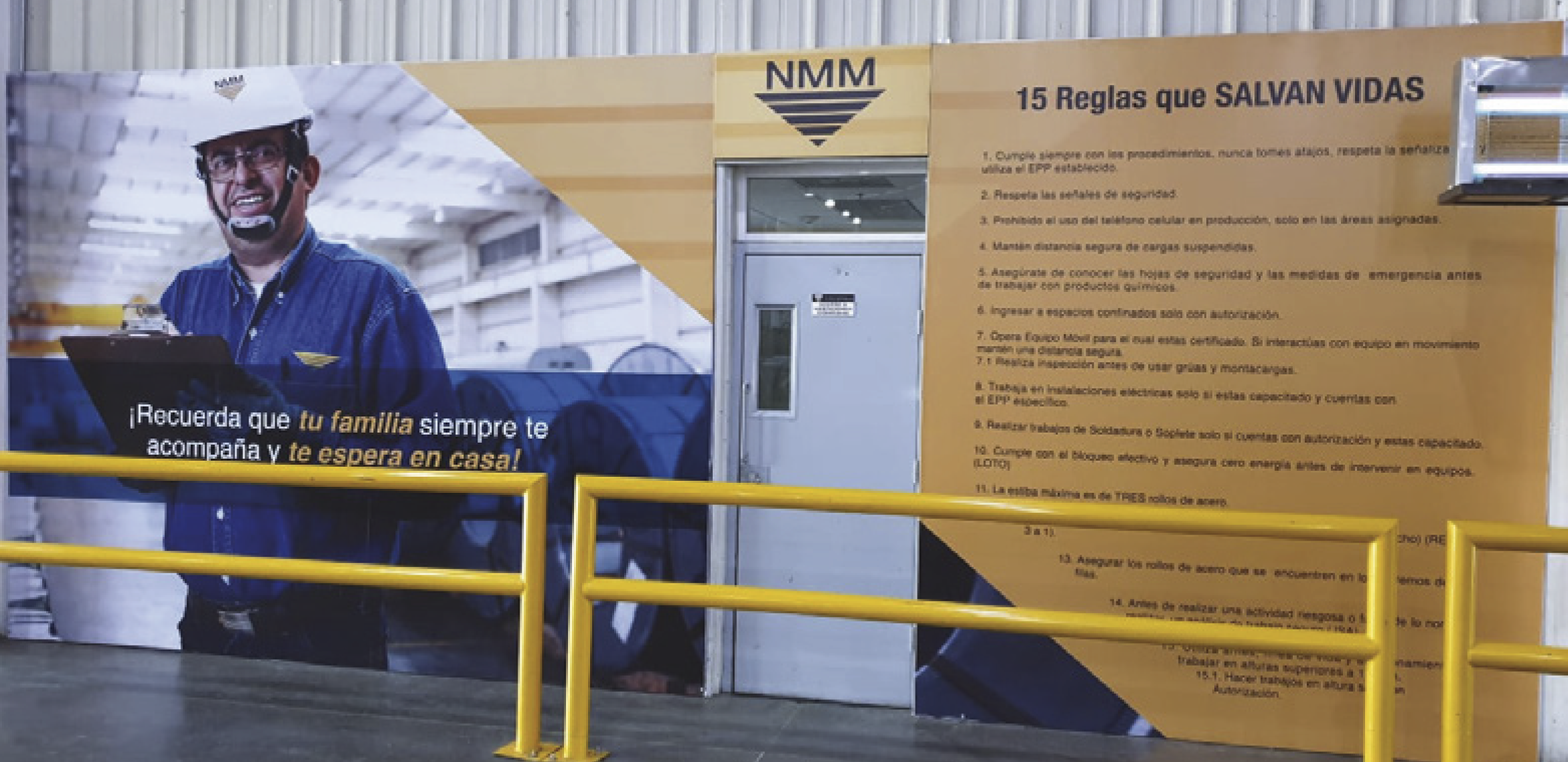 Sign at a workplace with a picture of a worker in blue uniform, wearing a safety helmet, glasses, and a headset, inside a large industrial facility. The sign includes the logo 'NMM' and text in Spanish listing 15 safety rules that save lives, along w