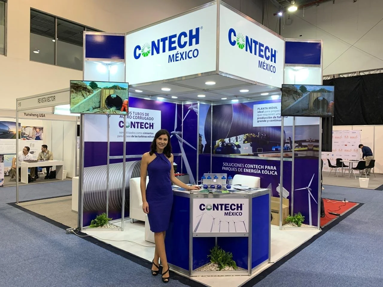 A woman in a blue dress standing at a trade show booth for Contech Mexico, which displays wind turbine components, including large pipes and a model wind turbine. The booth has purple and white branding with informational posters and screens showing 