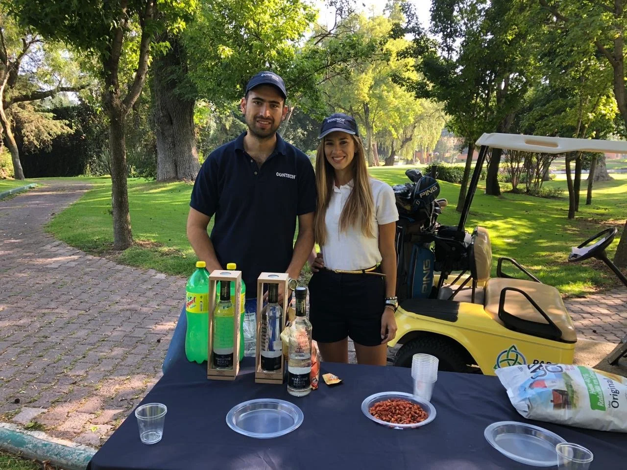 Two people standing behind a table with drinks and snacks on a golf course, with trees and a golf cart in the background.