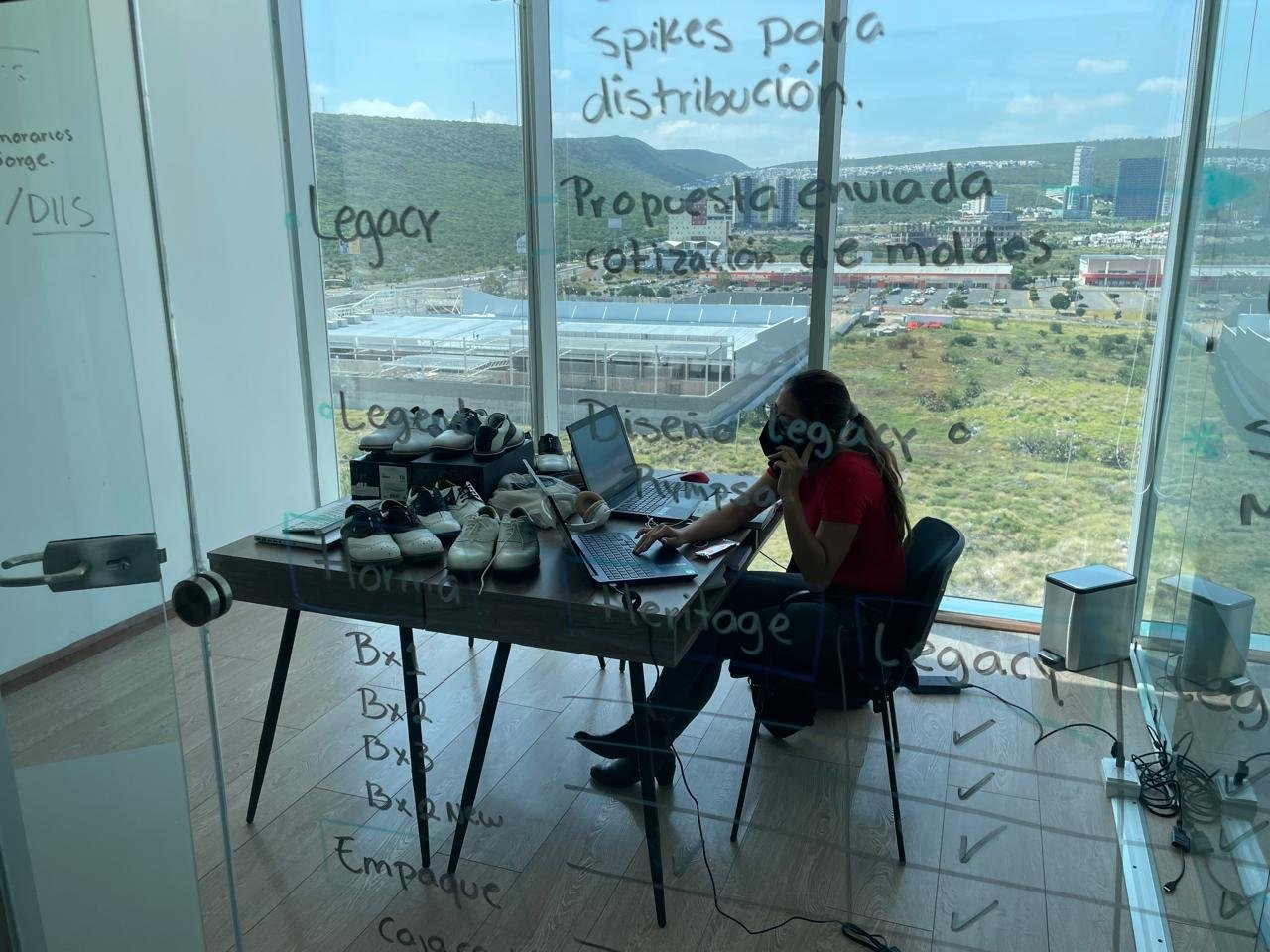 A woman in a red shirt working at a desk with multiple white shoes, laptop, and notebooks, inside a glass-walled office with a view of a city and green hills outside.