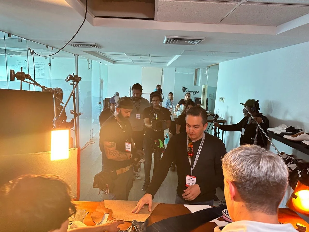 People gathered in a modern office or showroom, some wearing name tags, with a table, camera equipment, and shelves with shoes. The environment appears to be a professional event or product launch.