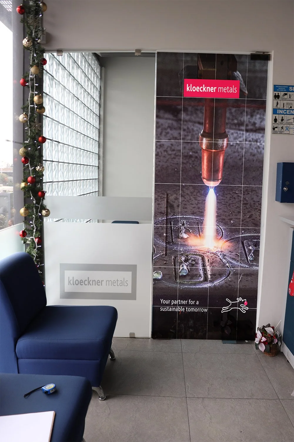 Indoor waiting area with a blue couch, Christmas decoration on the left, and a large Kloeckner Metals banner featuring a high-temperature metalworking process. The banner shows a torch melting metal with the text "kloeckner metals" and "Your partner 