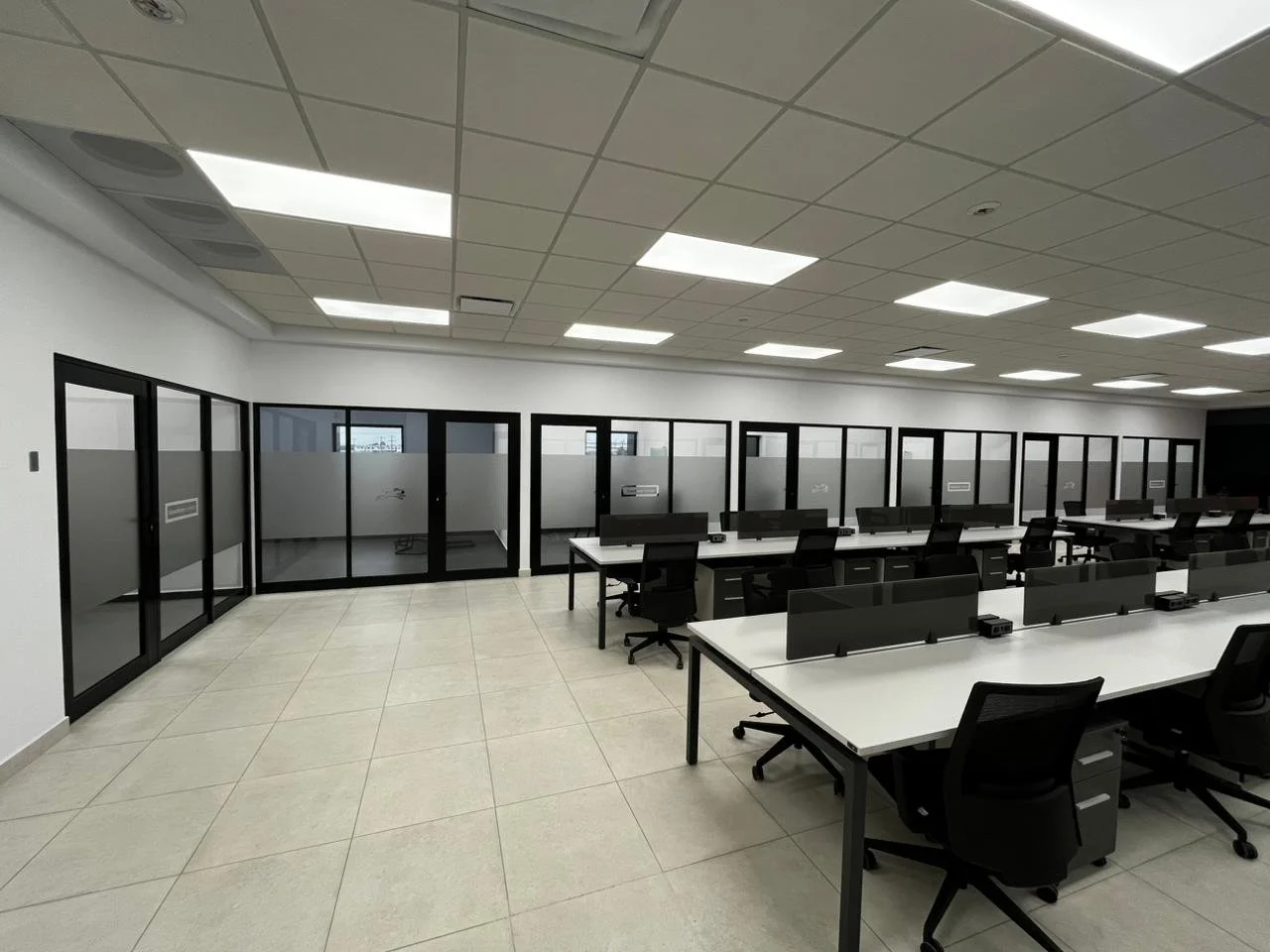 Empty office with desks, chairs, glass partition walls, tiled floor, ceiling lights, and white walls.