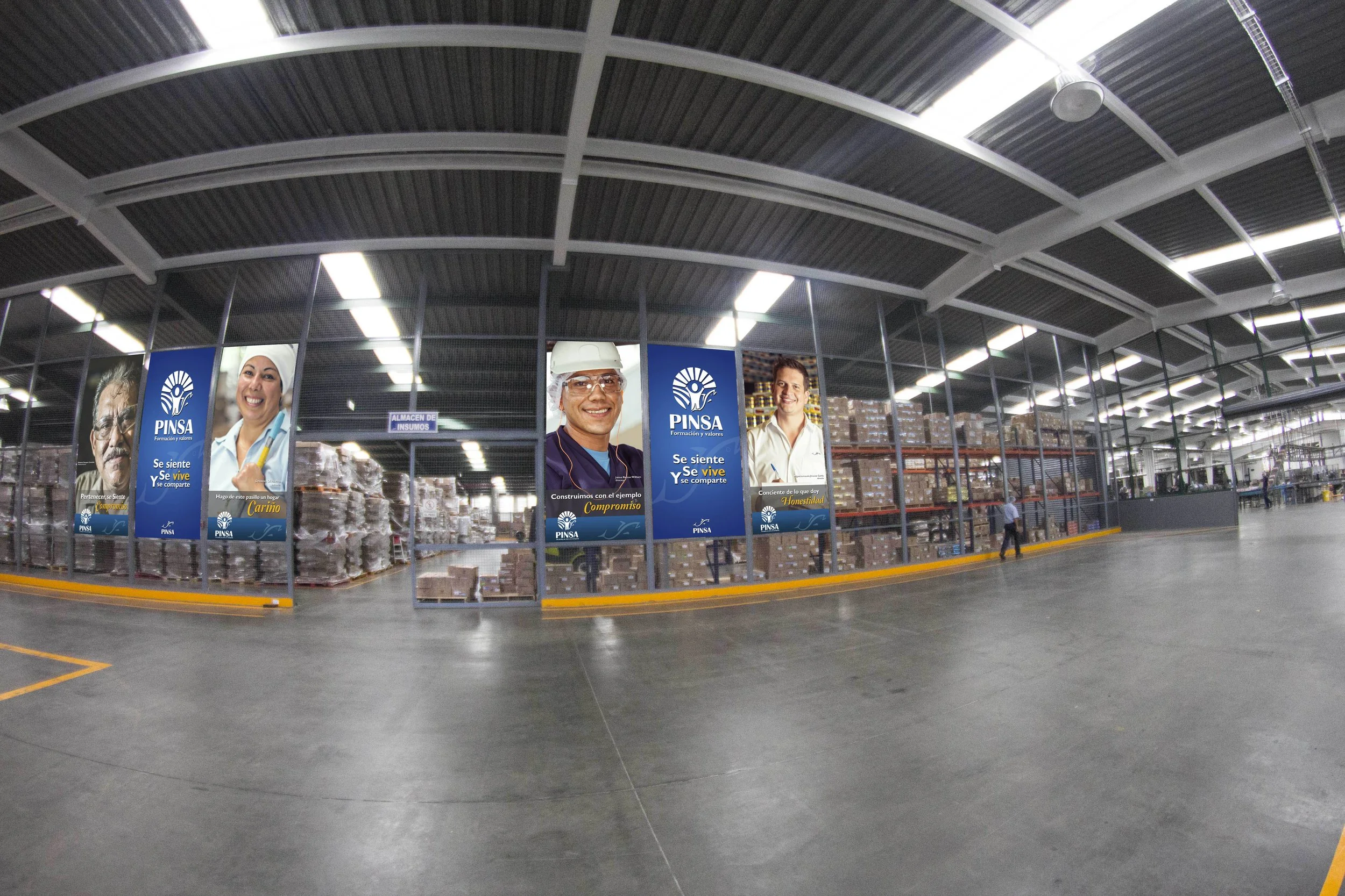 Inside a large warehouse with high ceilings and fluorescent lighting, shelves filled with boxes and pallets are visible. Large posters on the glass wall feature smiling people in work uniforms, promoting a company called PINSA with slogans encouragin