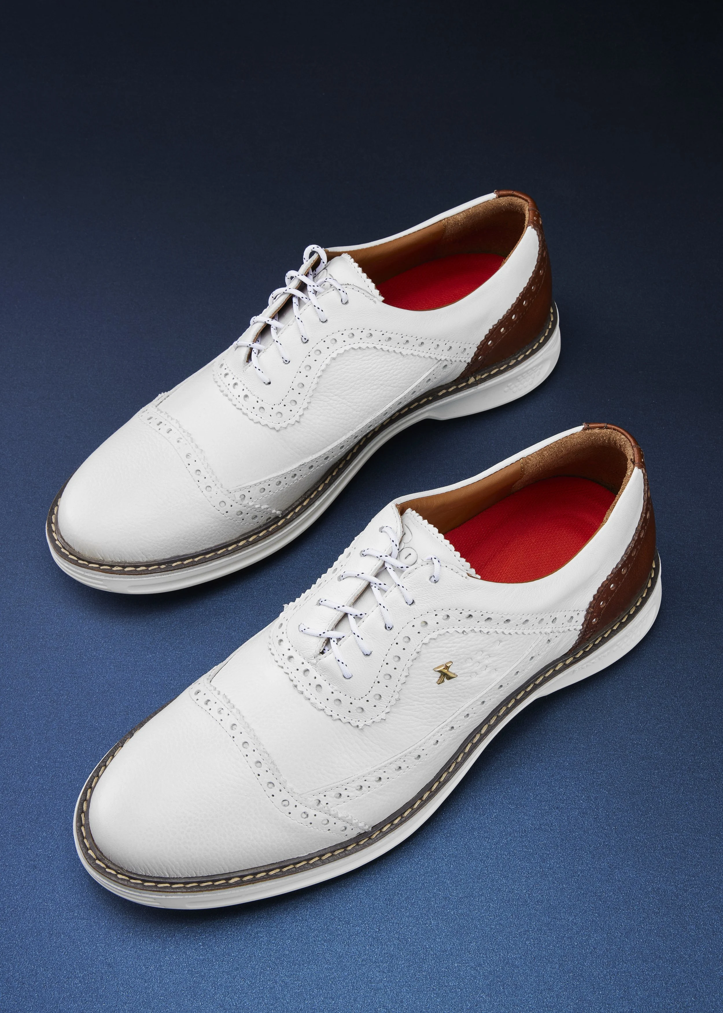 Pair of white leather brogue shoes with brown accents and red insoles placed on a dark blue surface.
