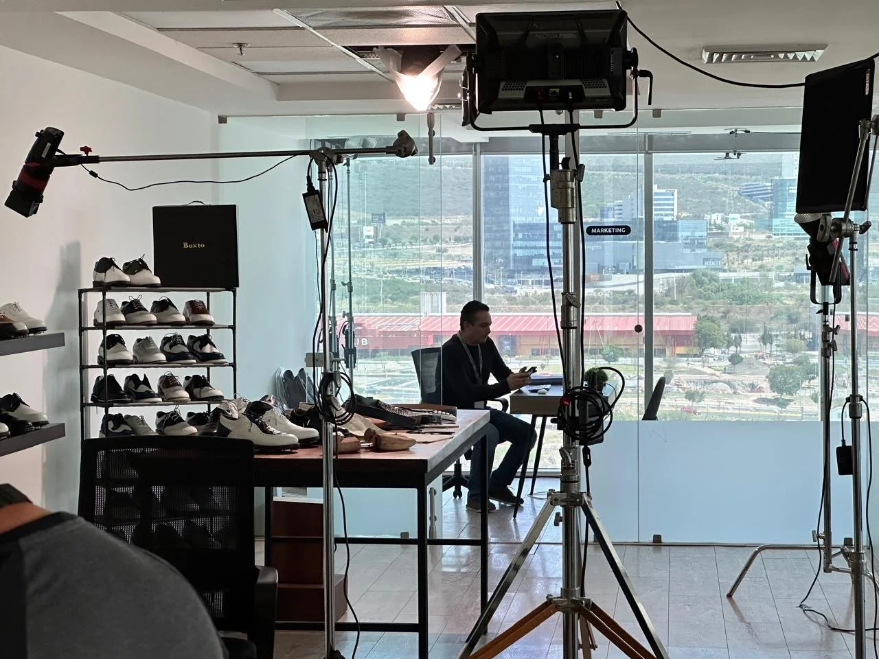 Interior of a modern office with a TV studio setup, a man sitting by the window using his phone, shelves with shoes, and filming equipment including lights, cameras, and microphones.