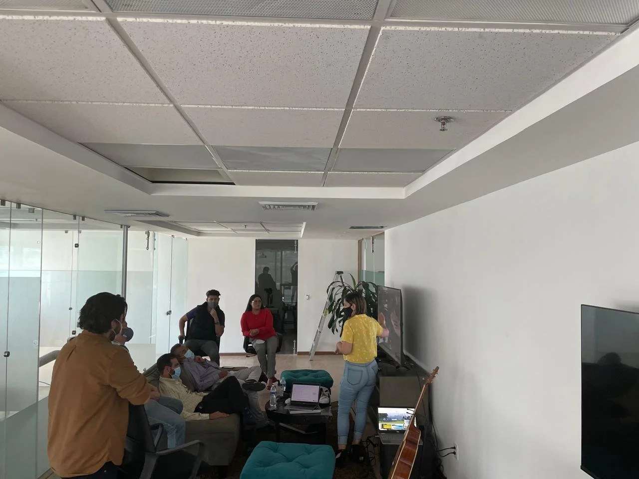 A group of people in a modern office or meeting room, some sitting on a couch and chairs, others standing or walking, with a woman in a yellow shirt addressing the group, a large TV screen on the wall, and a guitar leaning against the wall.