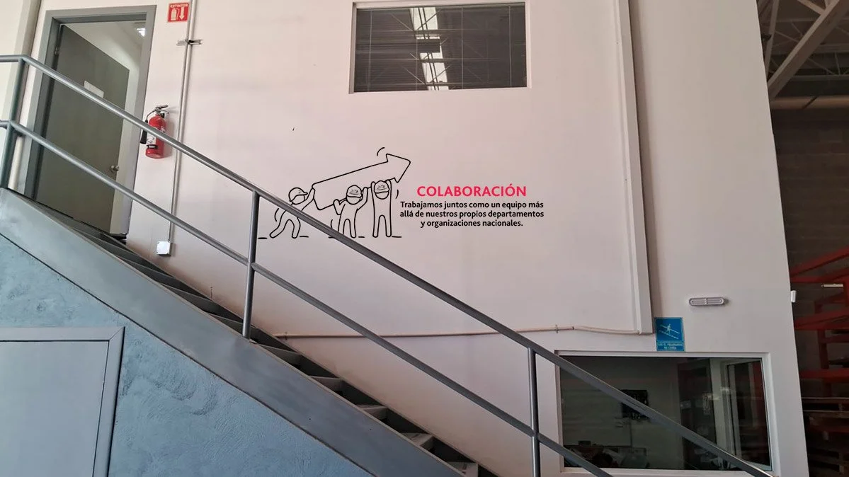 Interior of a building with a staircase and a white wall featuring a mural and text. The mural shows a simple line drawing of four people holding a banner together. The red text on the wall says 'COLABORACIÓN', and there is a paragraph explaining tea
