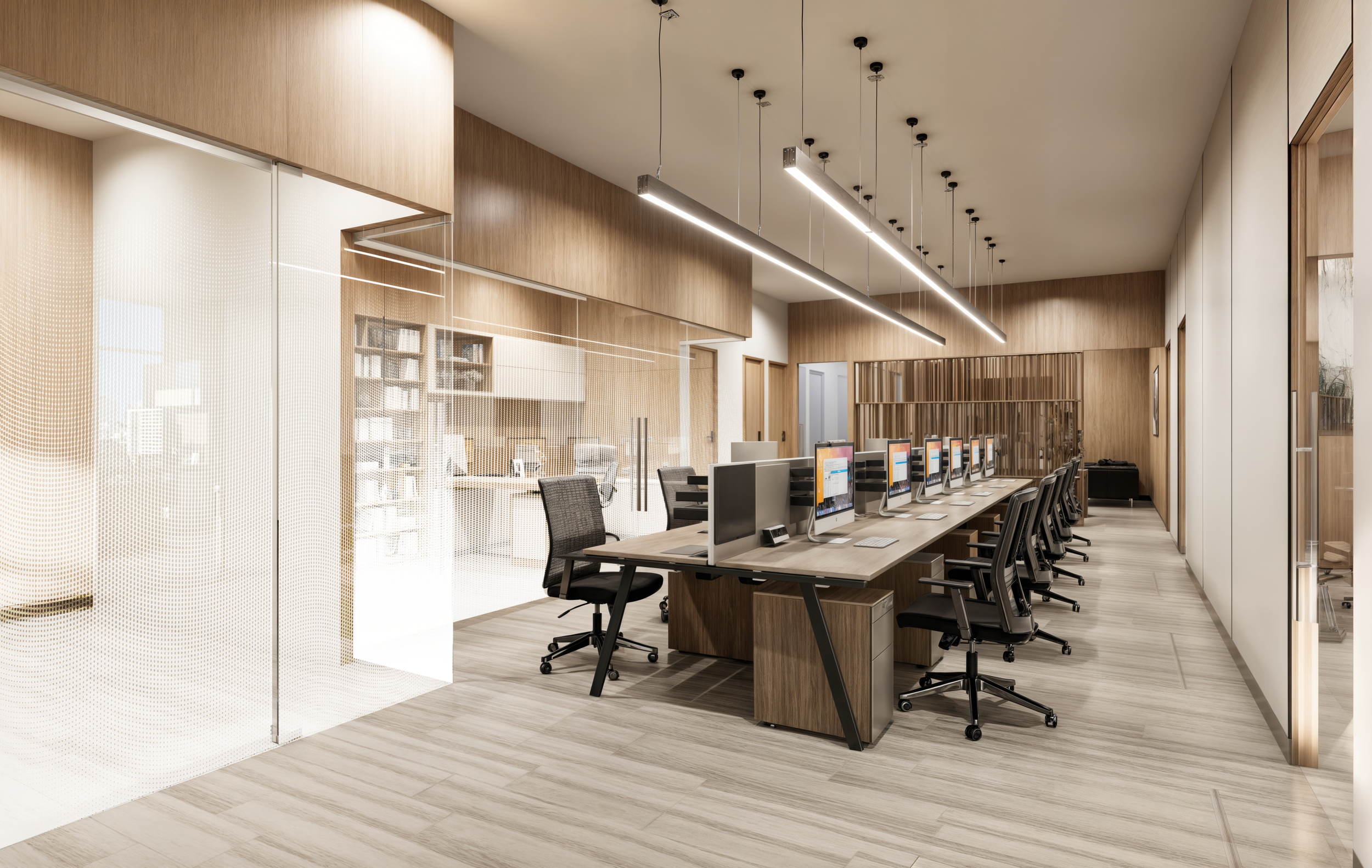 Modern office space with a row of computers and black office chairs on a wooden floor. The room has wood-paneled walls, glass partitions, and linear pendant lighting fixtures.