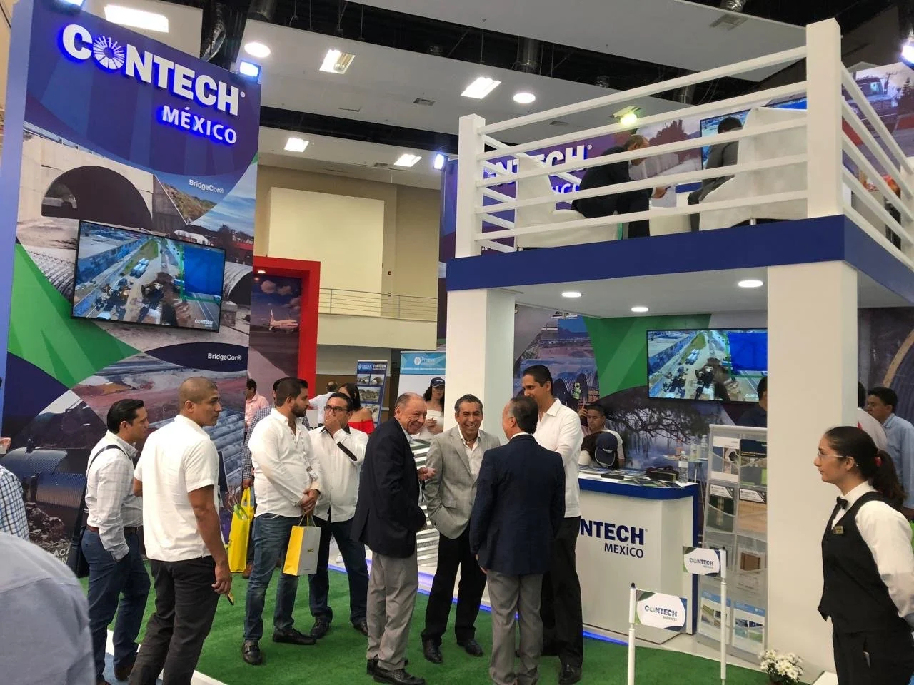 Trade show booth for CONTECH MEXICO with multiple people engaging in conversation. The booth features large screens showing construction or infrastructure projects, with promotional materials and a small seated area on an upper level. The setting is 