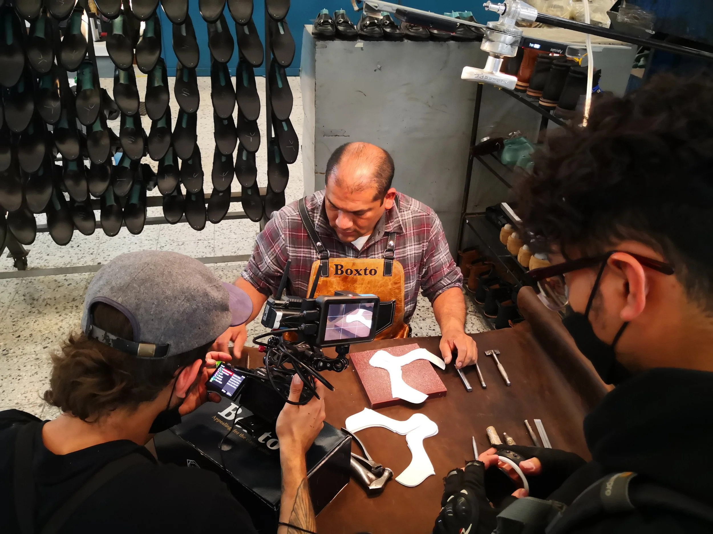 A man wearing an apron with the name 'Boxto' is demonstrating or explaining something with shoe parts on a table, while two people film or photograph him. The table has various tools and shoe components, and shoes are displayed on shelves in the back