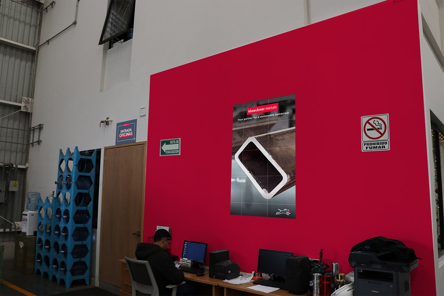 Office reception area with a red wall, poster, water dispenser, and person working at a desk with computers and office supplies.