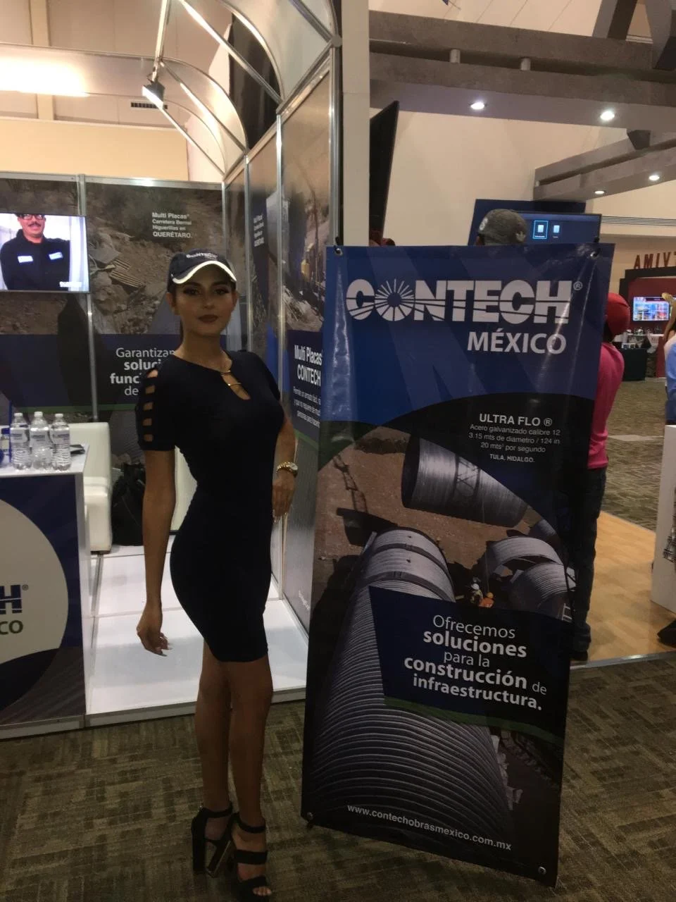 A woman in a black dress and high heels standing next to a Contech México banner at an indoor event, with people in the background.
