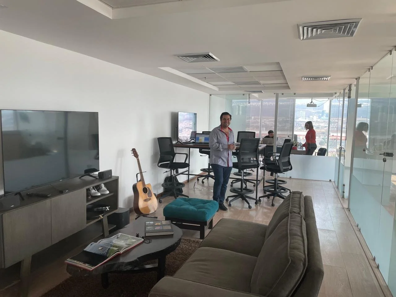 Modern office with large glass windows, a man standing in the center near a high table with chairs, a guitar leaning against the wall, a TV, a sofa, and a coffee table with items, two women working at a table by the window.