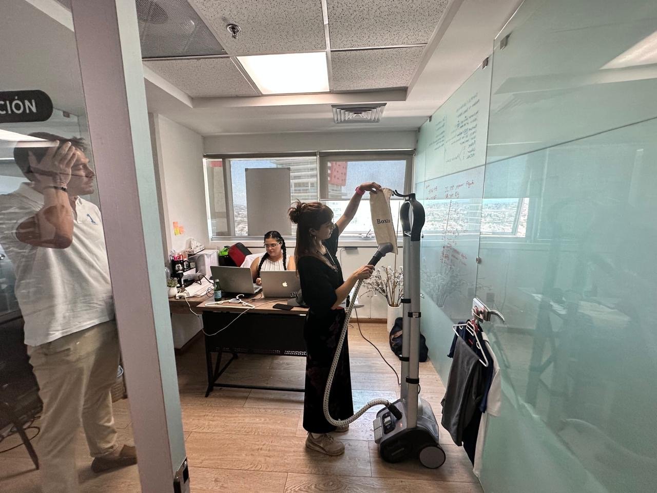 A woman is drying clothes with a vacuum cleaner in an office room, while two other women work on laptops at a desk, and a man looks on from the side.