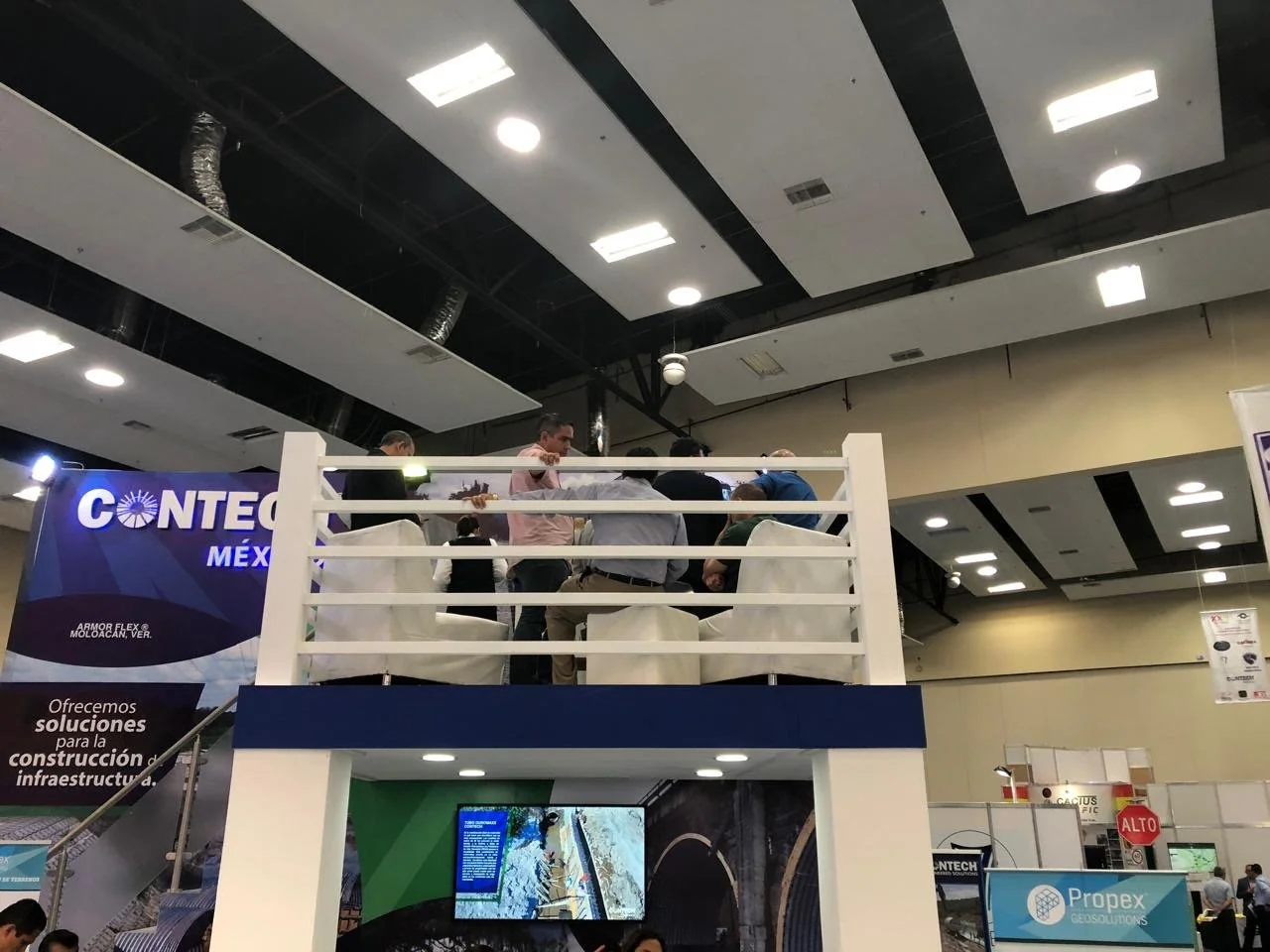 People gathered on a two-story exhibition booth at a conference or trade show, with banners displaying company information and a large monitor showing images.