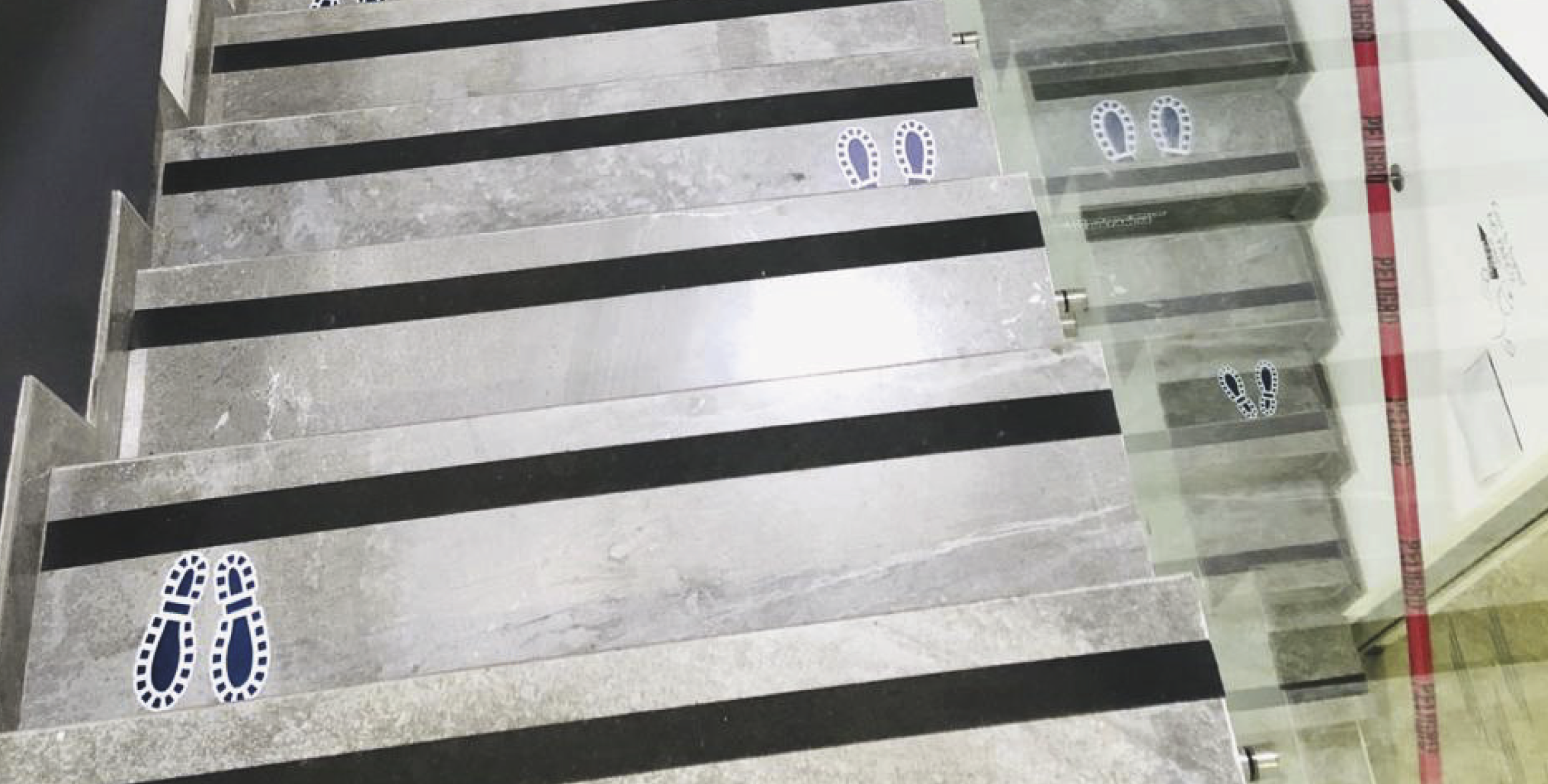 Floor markings with black stripes and blue social distancing footprints on a tiled surface, near a glass door with red warning tape.