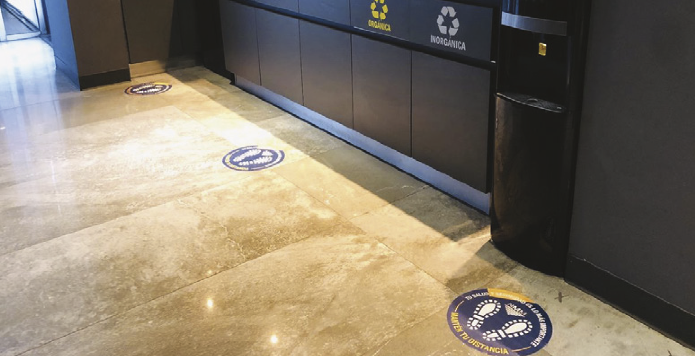 Floor stickers with social distancing instructions in a shopping mall or airport, advising people to maintain distance. The stickers have footprints and text in Spanish.