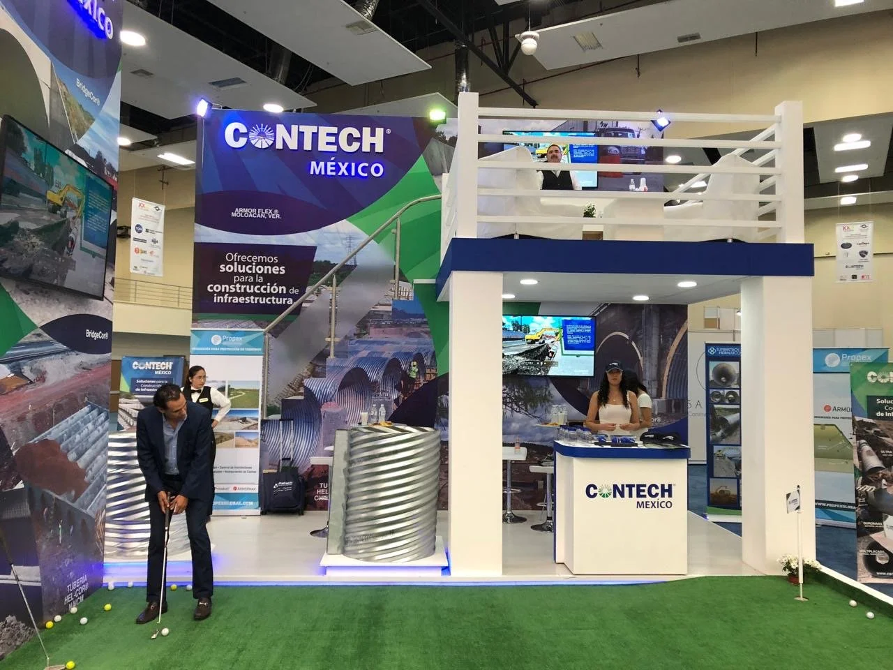 Trade show booth for CONTECH México featuring construction solutions and infrastructure projects. The booth has a two-story structure with banners, informational posters, a reception desk, and a man practicing golf on a small indoor putting green.