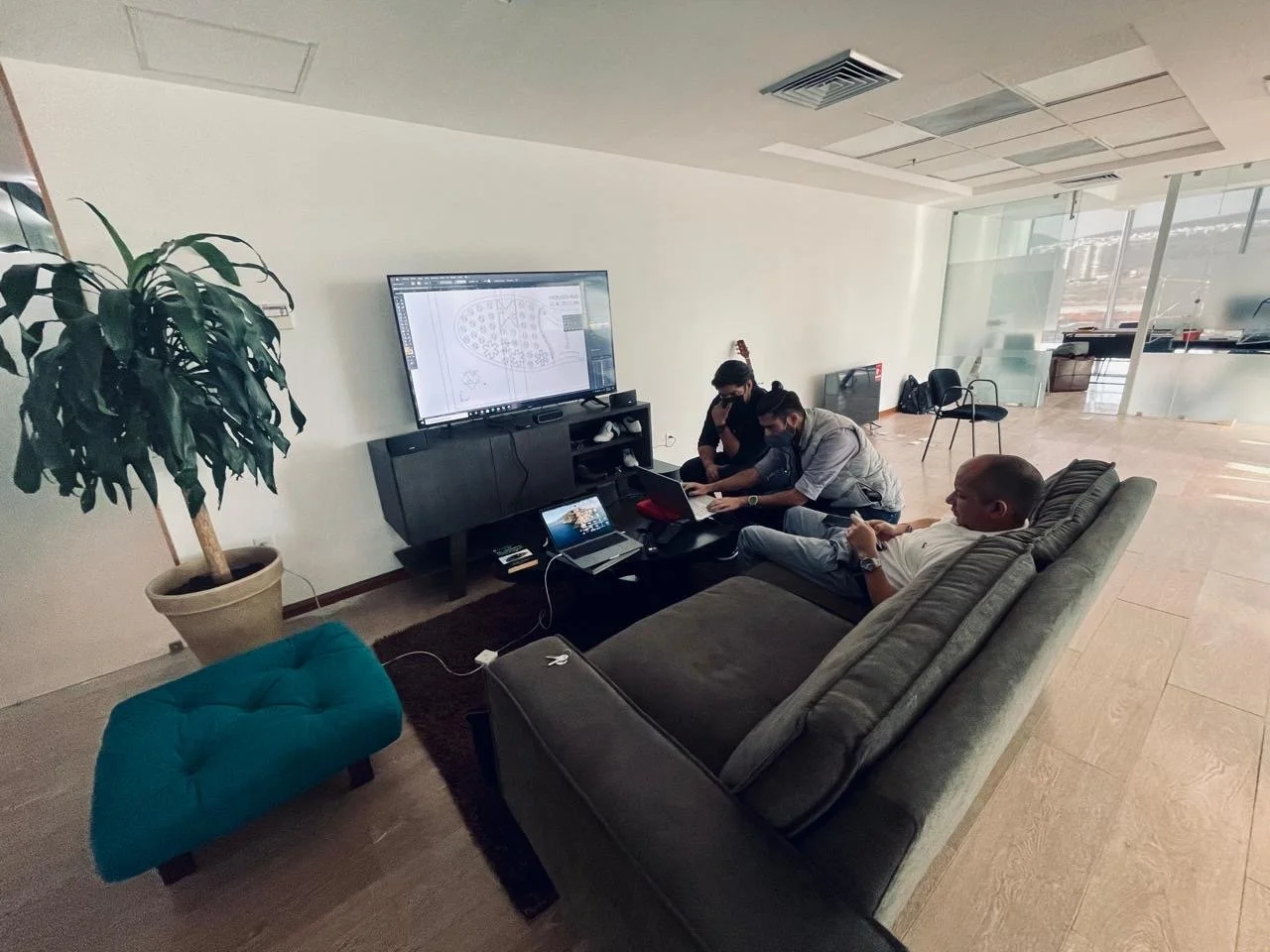 Three men sitting in a lounge area, two working on laptops and one using a smartphone, facing a large entertainment center with a television displaying a design software, next to a potted plant, with glass walls and chairs in the background.