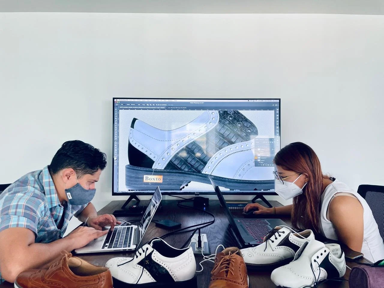 Two designers, wearing masks, working on sneakers at a table with laptops and digital design software displayed on a large screen, surrounded by various shoe samples.