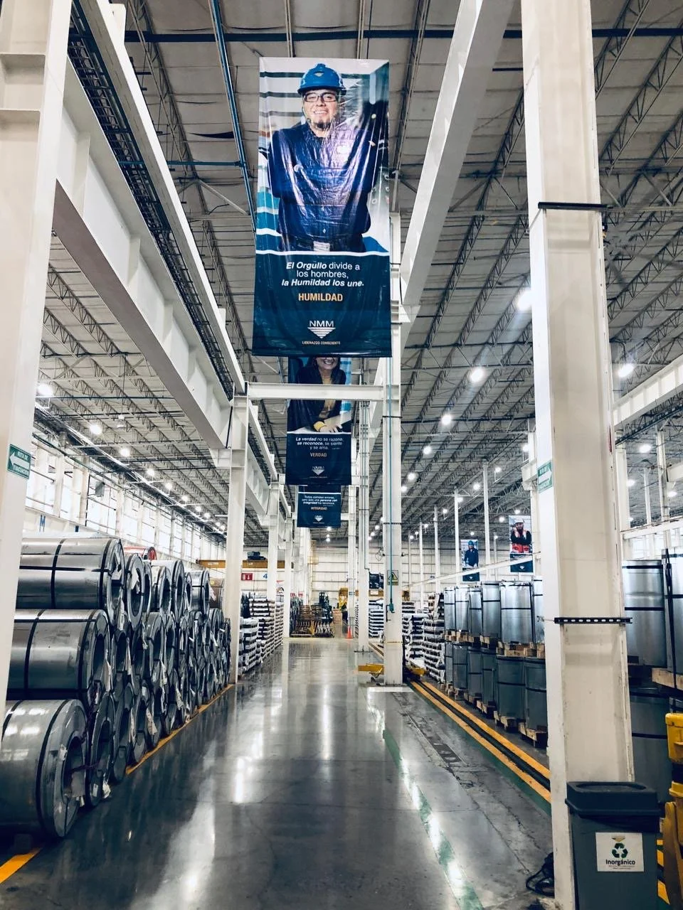 Industrial warehouse with metal coils and storage containers, hanging banners with a man wearing a construction helmet, high ceilings, and bright lighting.