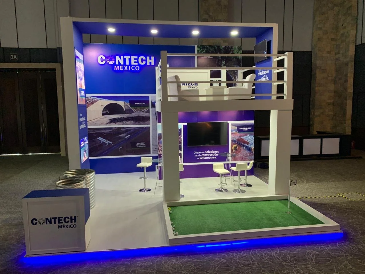 Exhibition booth for Contech Mexico with a two-story setup. The booth features a large purple wall with the Contech Mexico logo, a television screen, white chairs, and a small green artificial turf area with a golf club and golf ball. There are also 