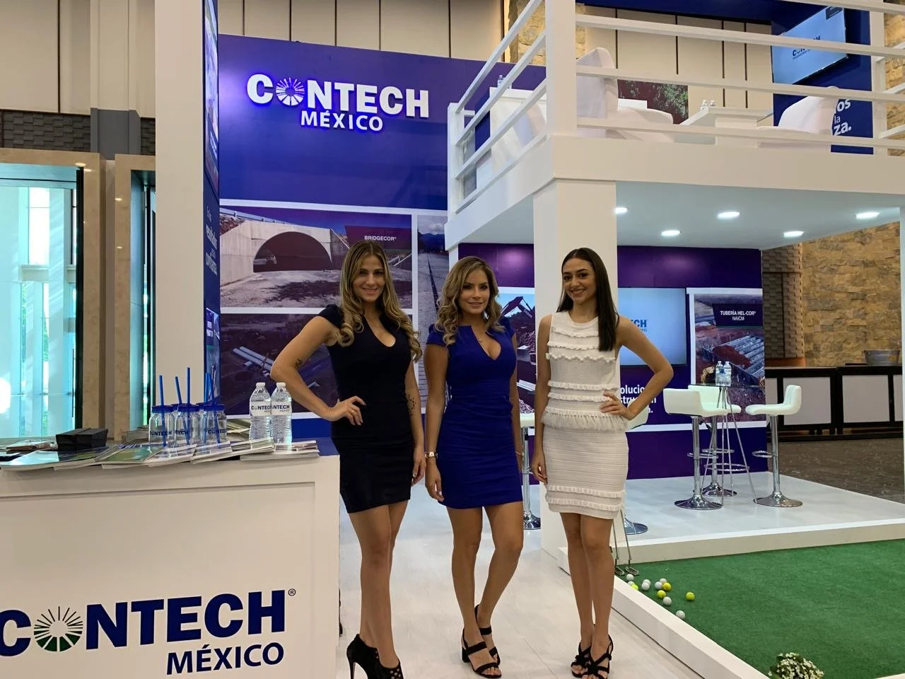 Three women standing in front of a CONTECH Mexico exhibition booth at a trade show, with informational displays and green artificial turf with golf balls to the side.