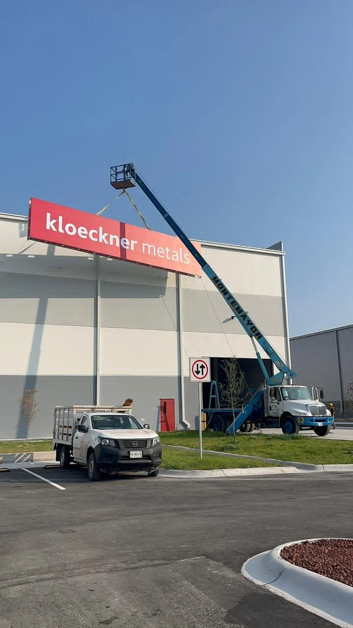 A crane lifting a sign that reads 'kloeckner metals' onto a building. There is a white truck parked in front and a no parking sign nearby. The setting appears to be a commercial or industrial area.