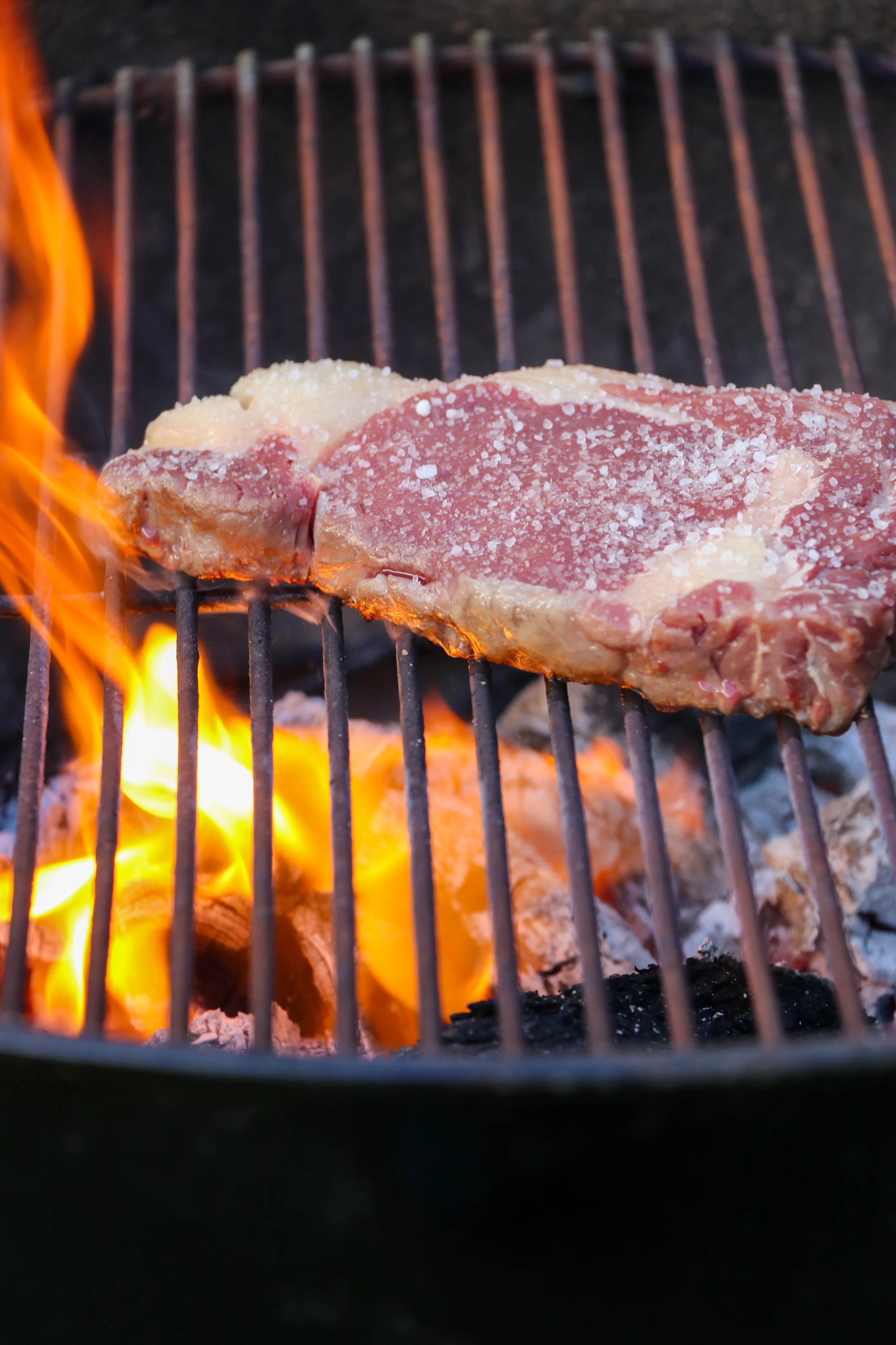 A piece of seasoned uncooked steak on a grill over open flames.