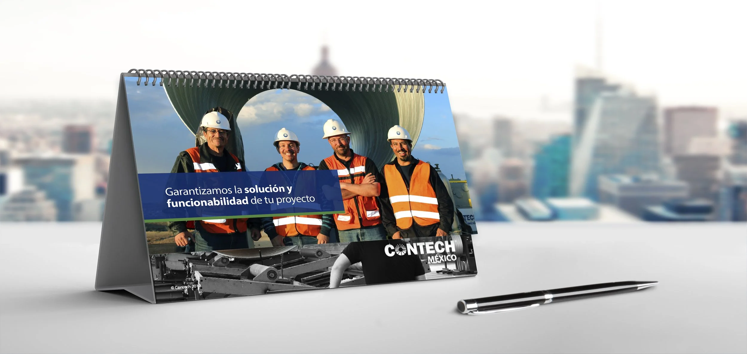 A desk calendar with a photo of four construction workers in safety vests and helmets, standing in front of a large industrial pipe, with a city skyline in the background. The calendar contains text in Spanish that translates to "We guarantee the sol