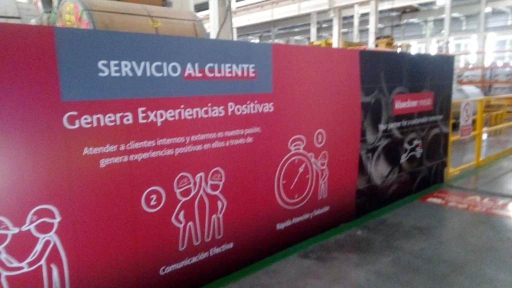 Sign in a warehouse or industrial setting with red, blue, and black panels. The sign reads 'Servicio al Cliente' and 'Genera Experiencias Positivas,' with icons depicting communication and time management.