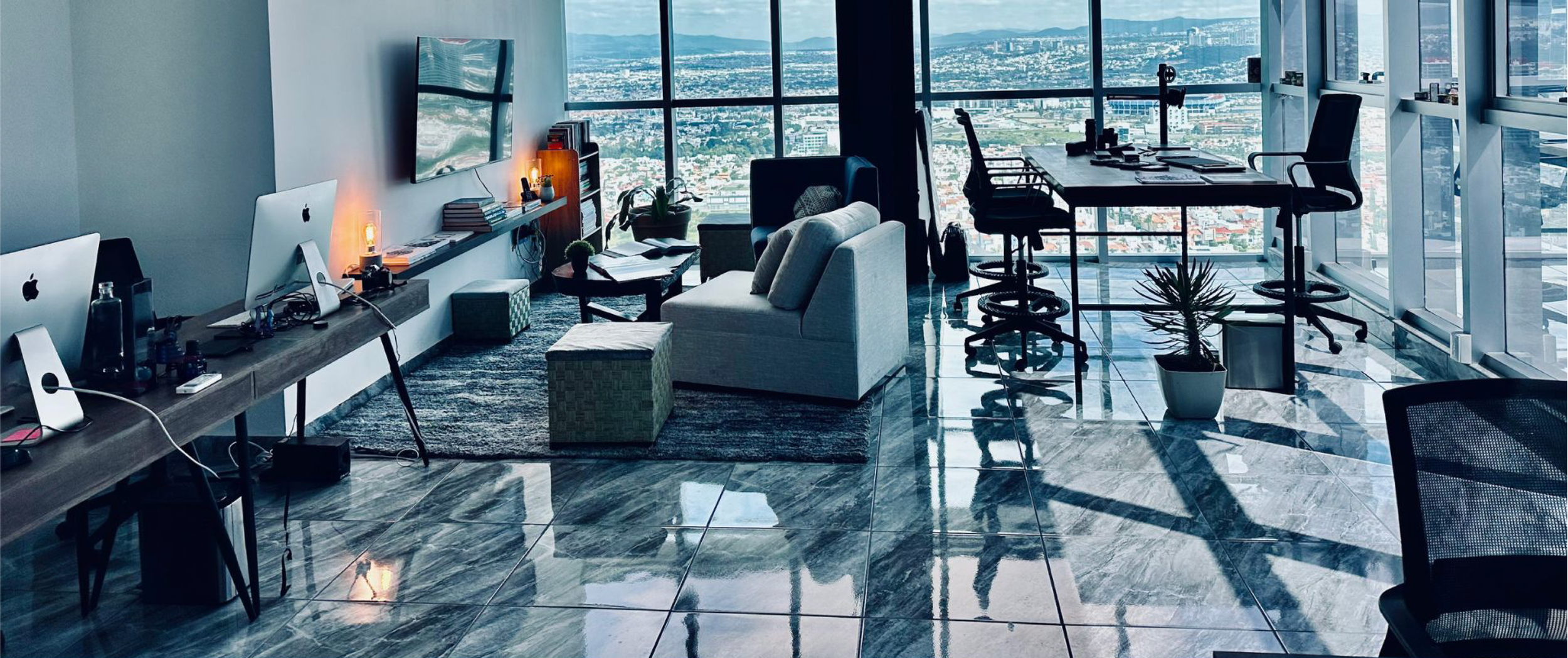 Modern office with large windows overlooking cityscape, featuring a desk with computers, a cozy sitting area with armchair and ottoman, and a high table with stools, all illuminated by daylight.