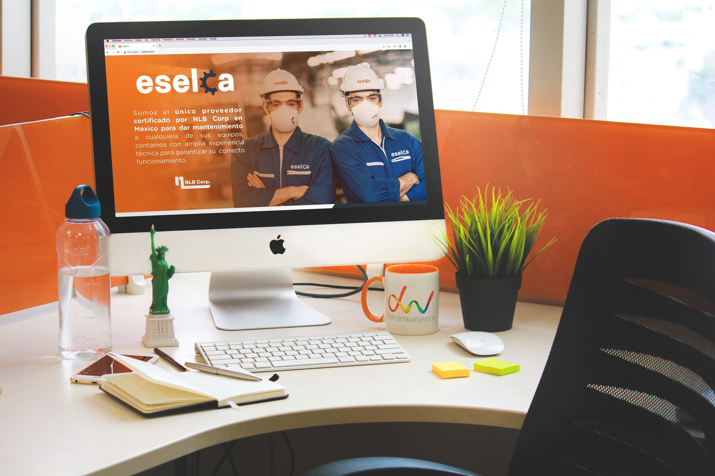 Office desk with an Apple iMac displaying a website for Eselca, featuring two workers in safety gear and masks. The desk has a water bottle, a miniature Statue of Liberty, a notebook with a pen, a Mug with a colorful logo, a potted plant, sticky note