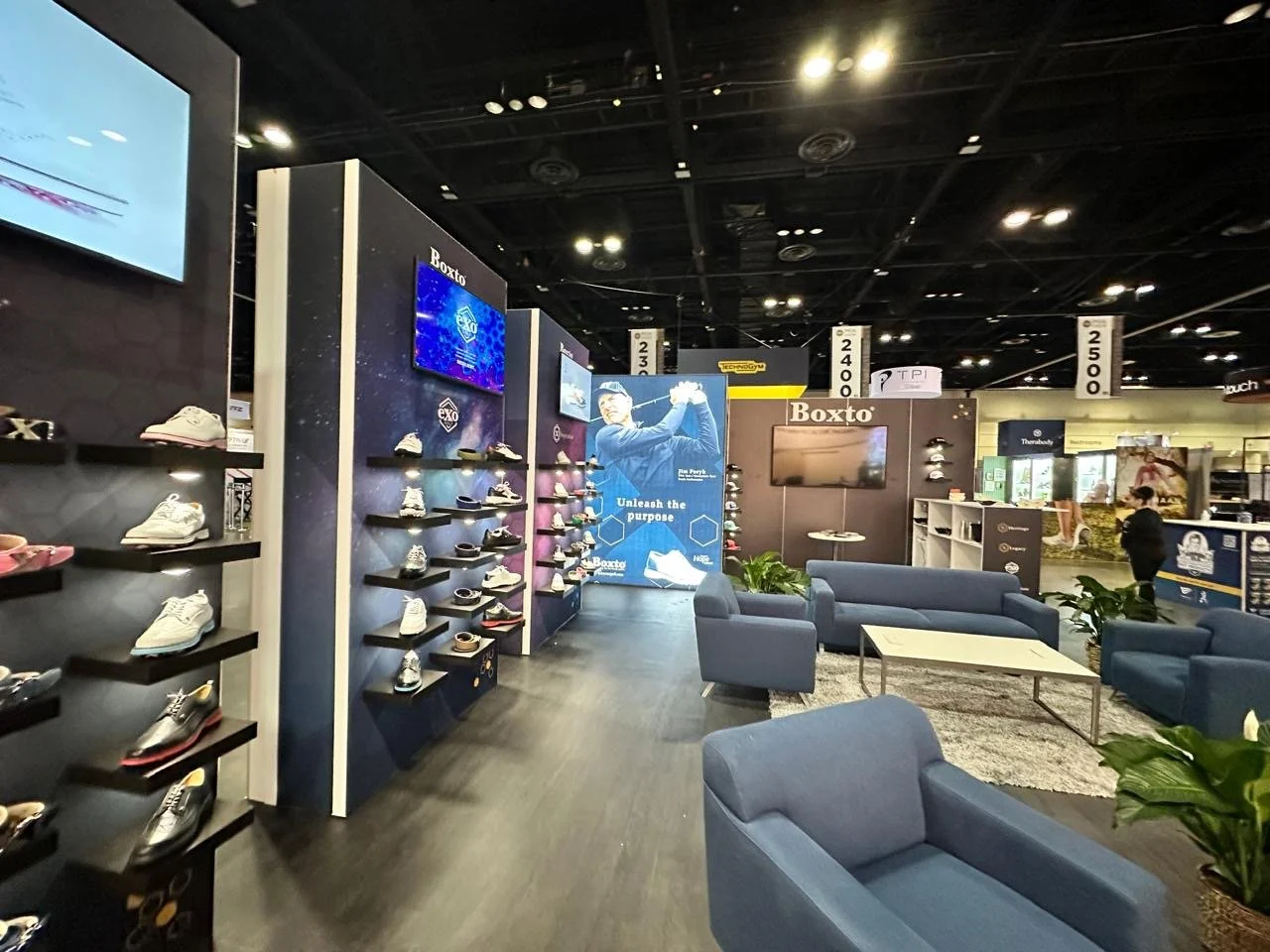 An indoor exhibition area with shoes displayed on black shelves along a wall, a seating area with blue sofas and a coffee table, and promotional banners including the brand name 'Boxto' and athletic footwear. The space features a dark ceiling with ex