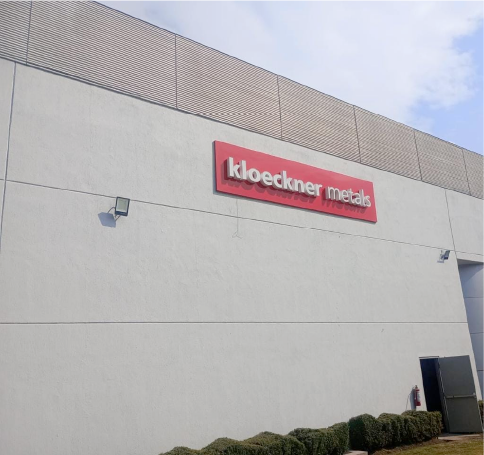 Kloeckner Metals building exterior with red sign, light gray walls, small bushes, and a black vent or air unit.
