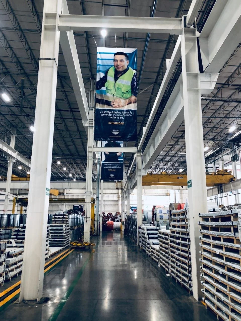 Interior of a large industrial warehouse with metal racks filled with coiled cables and equipment. Hanging banners with a smiling man in a safety vest and Spanish text.