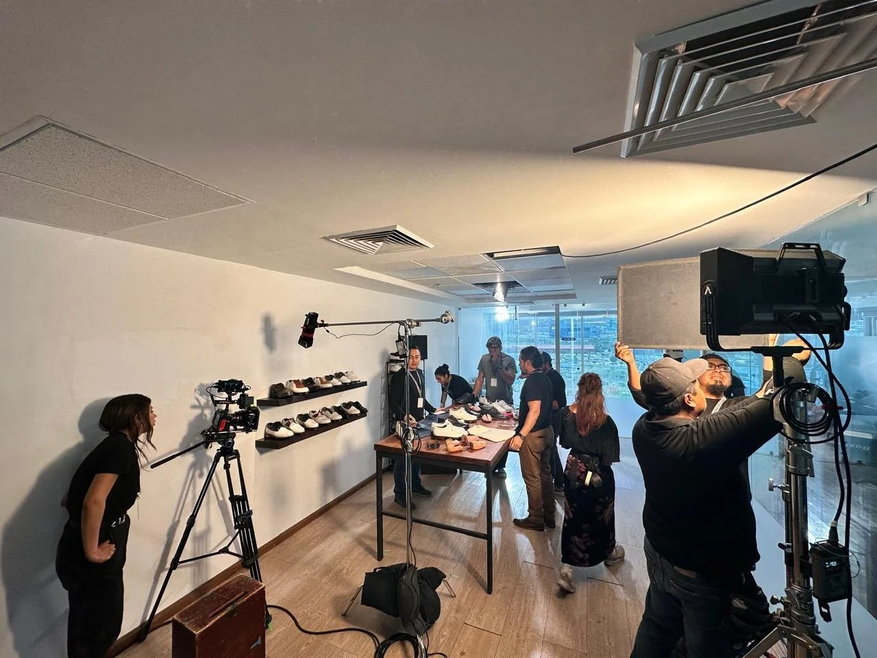 A filming crew set up a shoot in a room with people examining shoes on a table. Camera equipment and lighting are visible, and a camera operator adjusts a light on a stand. The room has a large window with a cityscape view in the background.