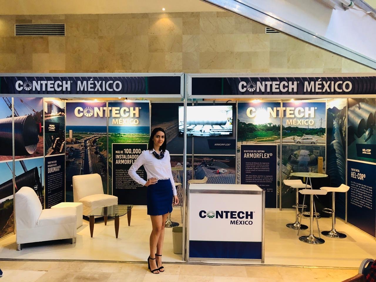 A woman in professional attire standing inside a CONTECH Mexico trade show booth with informational posters, a small seating area, and high tables.
