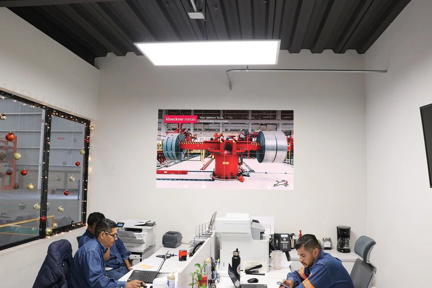 An office with three men working at desks, one using a laptop and another on a phone. The room has white walls, a black ceiling, and a large poster of industrial machinery on the wall. There is a window with holiday decorations on the left side of th