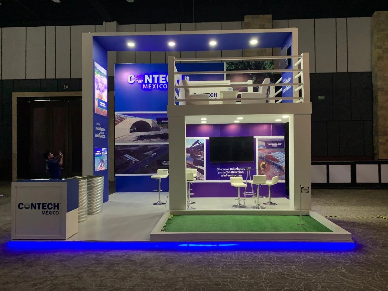 Trade show booth for CONTECH México with a two-story structure. The booth features blue and purple branding, a sitting area with chairs on the upper level, and a small green turf area on the lower level. There are informational displays and a large T