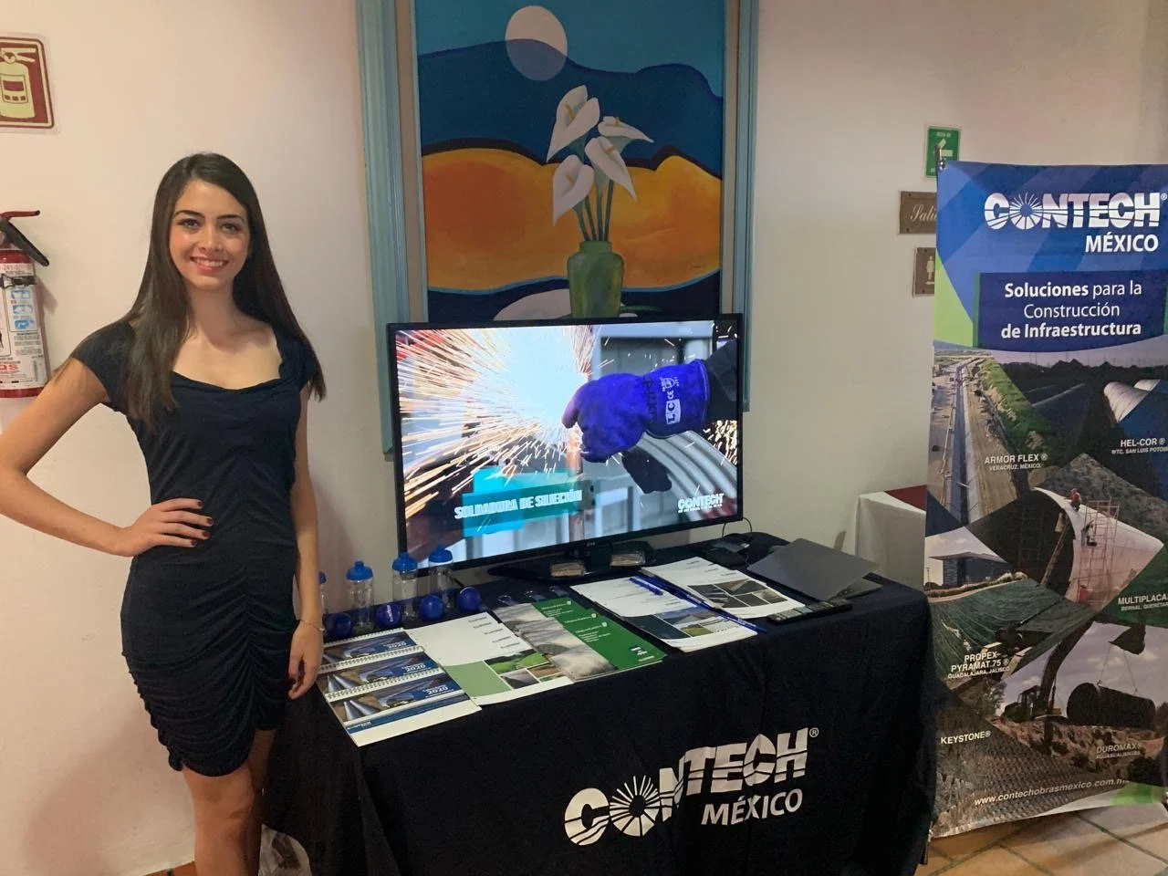 A young woman with long dark hair wearing a black dress stands next to a conference booth for Contech México. The booth features a table with brochures and a flat-screen TV displaying an image of a worker welding with sparks. Behind her is a colorful