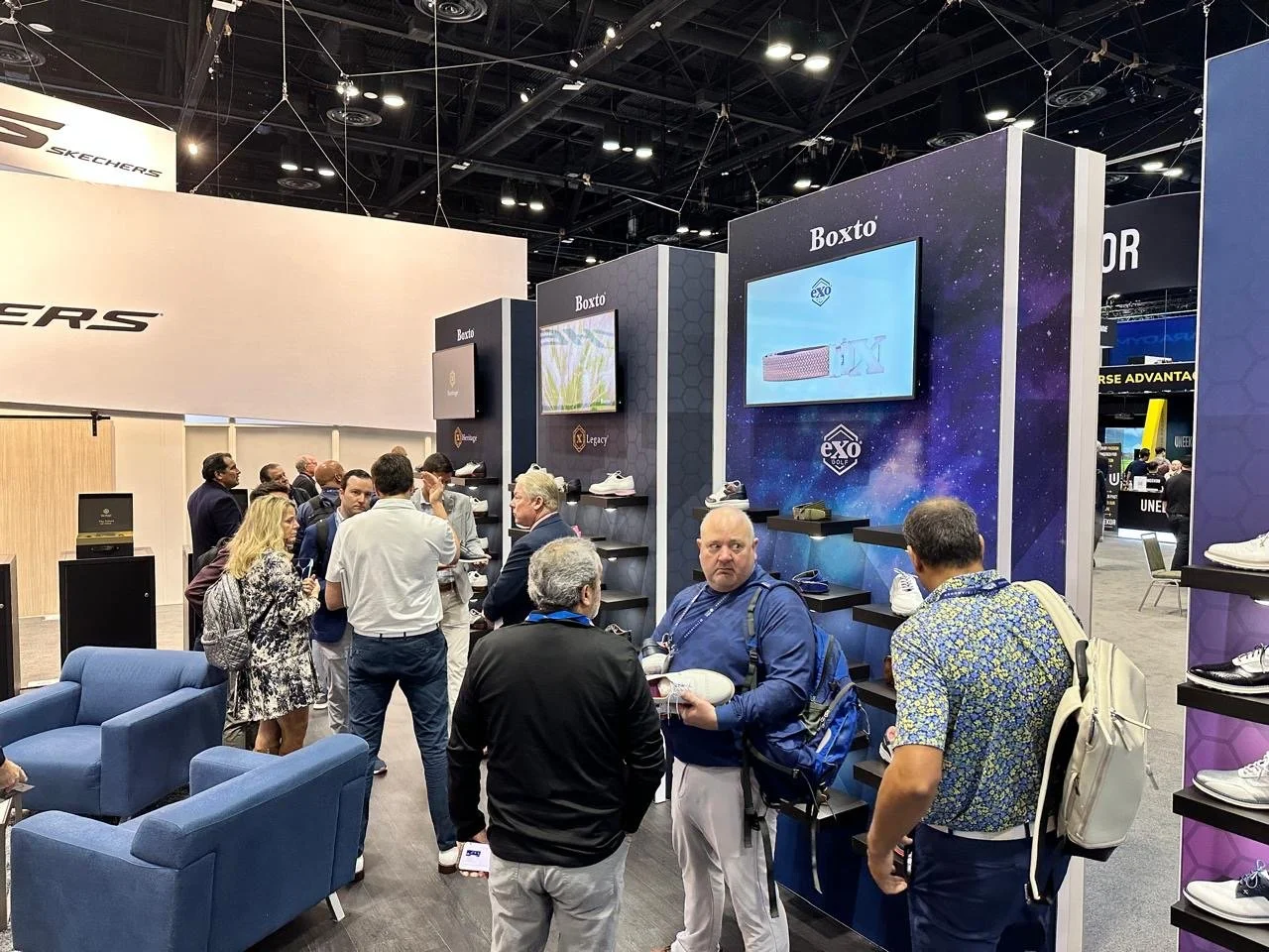 People at a sneaker booth in a convention center, with shelves displaying various sneakers and large screens showcasing shoe images and logos.